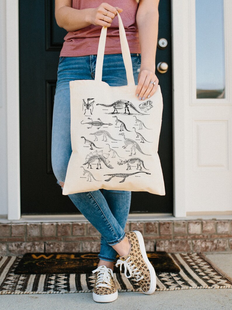 May include: A beige tote bag with a black and white illustration of dinosaur skeletons. The bag has two handles and is being held by a person wearing blue jeans.