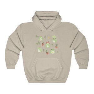 May include: A light beige hoodie with a pattern of green frogs, plants, and mushrooms. The frogs are wearing crowns, hats, and holding flowers.