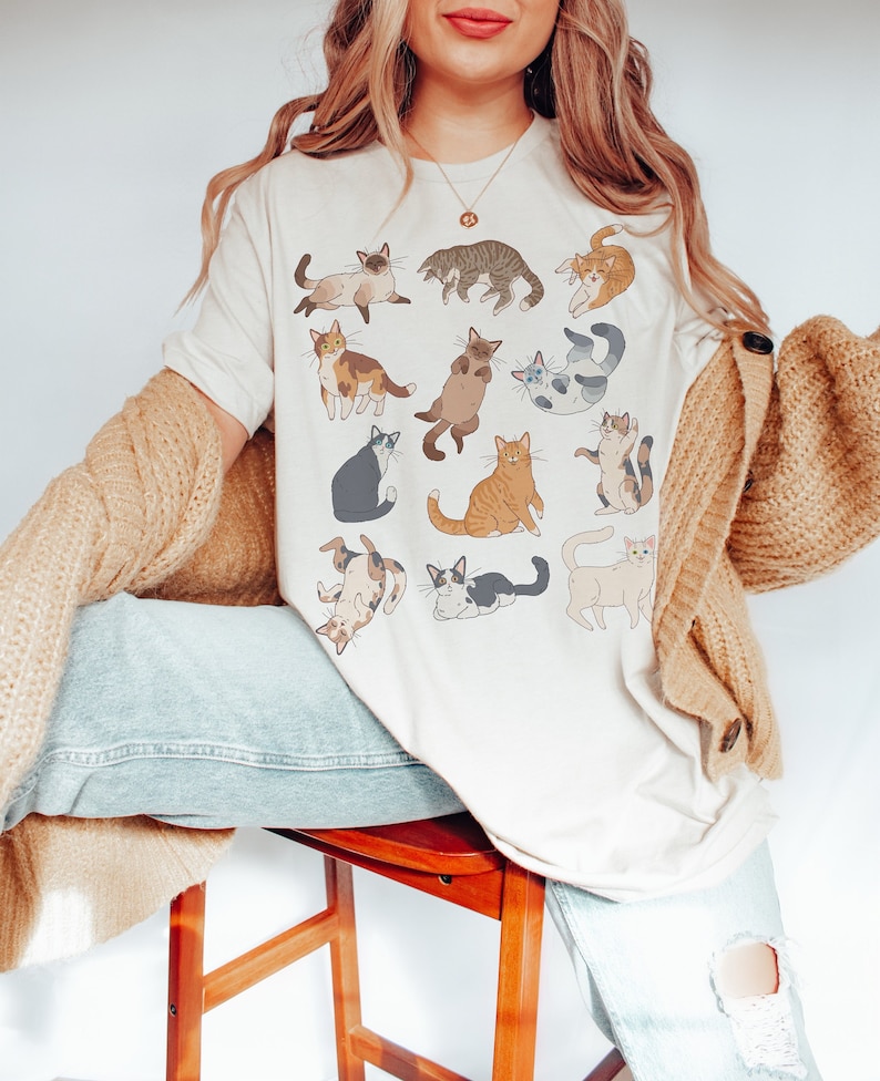 May include: Cream-colored t-shirt featuring a collection of illustrated cats in various poses. The cats are depicted in a range of colors, including brown, gray, orange, and white. The shirt is a casual, comfortable style.