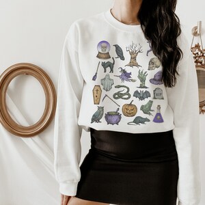 May include: White sweatshirt with a black and white graphic print of various Halloween icons, including a skull, a ghost, a witch, a cat, a snake, a cauldron, a pumpkin, a bat, a frog, a potion bottle, a crow, a tree, a coffin, a grave, a scythe, a wand, and a crystal ball.