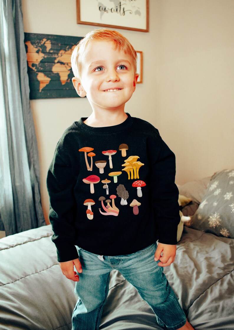 May include: A young boy wearing a black sweatshirt with a colorful mushroom print and the text "Shroom Queens". He is wearing blue jeans and standing on a gray bed.