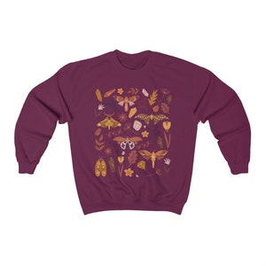 Puede incluir: Una sudadera color granate con un patr&oacute;n colorido de polillas, cristales y hojas. El dise&ntilde;o presenta una variedad de especies de polillas, incluyendo polillas luna, polillas halc&oacute;n y polillas esfinge.