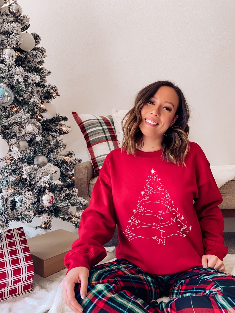 May include: A red Christmas sweatshirt featuring a white outline of a Christmas tree made of pigs. The person wearing the sweatshirt is sitting on a white fluffy surface wearing red and green plaid trousers.