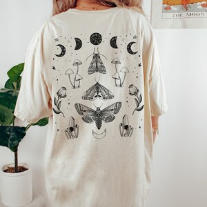 Op de afbeelding: Een cr&egrave;mekleurig T-shirt met een zwart-witte grafische print van motten, paddenstoelen, kristallen en halve manen. De afbeelding bevindt zich in het midden op de achterkant van het shirt.