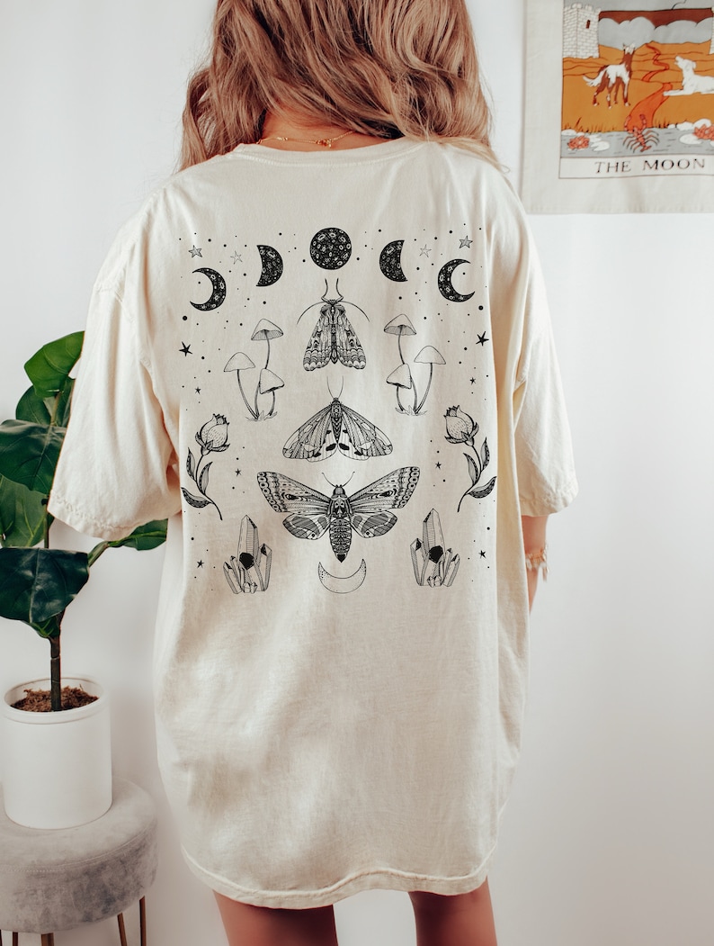 Op de afbeelding: Een cr&egrave;mekleurig T-shirt met een zwart-witte grafische print van motten, paddenstoelen, kristallen en halve manen. De afbeelding bevindt zich in het midden op de achterkant van het shirt.