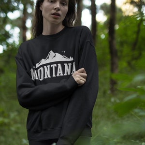 May include: A black sweatshirt with a white graphic of a mountain range and the word "MONTANA" in a vintage style font. The person wearing the sweatshirt is standing in a forest.
