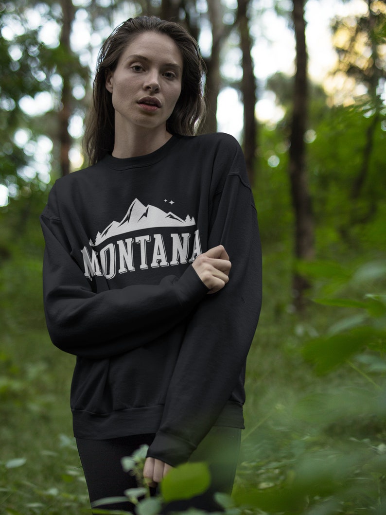 May include: A black sweatshirt with a white graphic of a mountain range and the word "MONTANA" in a vintage style font. The person wearing the sweatshirt is standing in a forest.