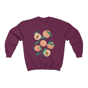 K&ouml;nnte beinhalten: Ein maroonfarbener Pullover mit einem Grafikdruck von Pfirsichen mit gr&uuml;nen Bl&auml;ttern. Die Pfirsiche sind in einem Kreis angeordnet.