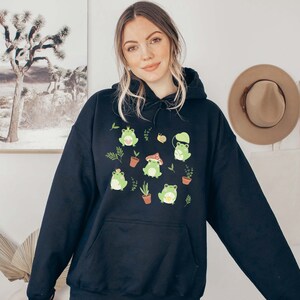 May include: Black hooded sweatshirt with a graphic print of green frogs wearing hats and holding mushrooms. The frogs are surrounded by small plants and flowers.