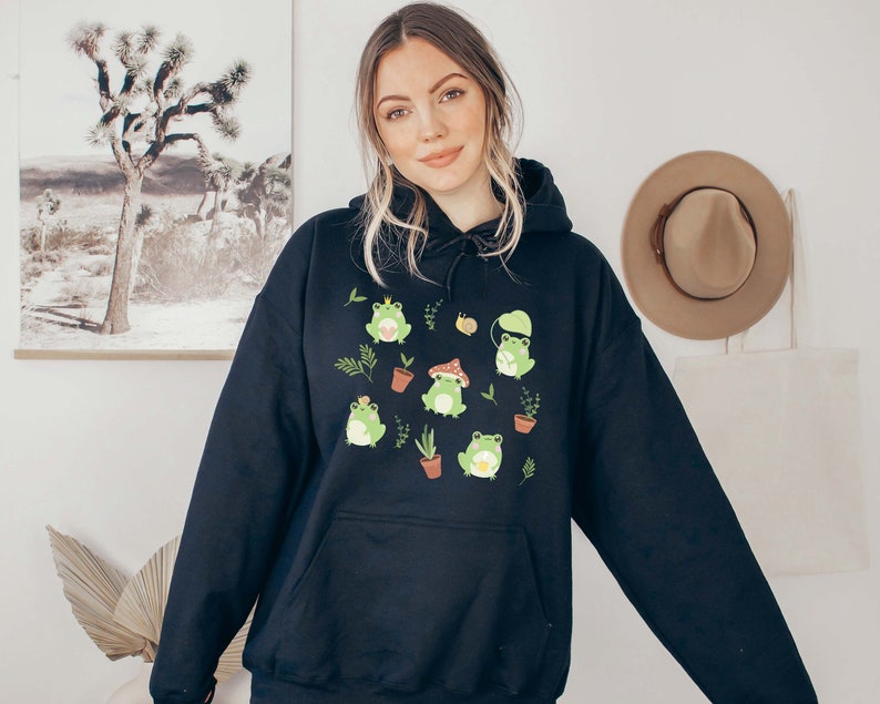 May include: Black hooded sweatshirt with a graphic print of green frogs wearing hats and holding mushrooms. The frogs are surrounded by small plants and flowers.