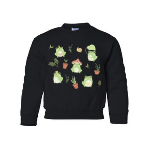 K&ouml;nnte beinhalten: Schwarzer Pullover mit einem Muster aus gr&uuml;nen Fr&ouml;schen und Pilzen. Die Fr&ouml;sche tragen Kronen und halten Blumen. Der Pullover hat einen Rundhalsausschnitt und lange &Auml;rmel.