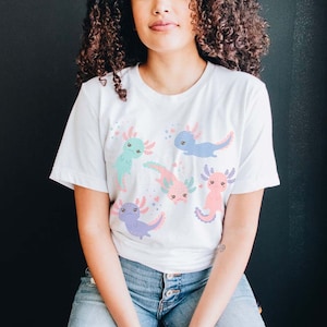 May include: White t-shirt featuring a colourful print of five axolotls. The axolotls are in various poses and have different coloured bodies and fins. The shirt has short sleeves and a crew neck.