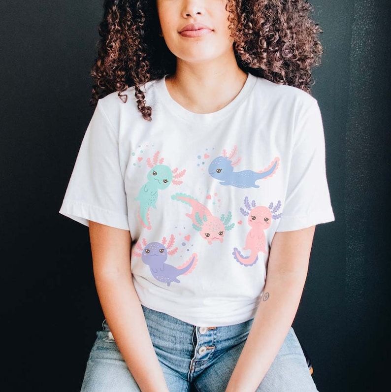 May include: White t-shirt featuring a colourful print of five axolotls. The axolotls are in various poses and have different coloured bodies and fins. The shirt has short sleeves and a crew neck.