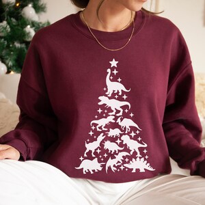 May include: A maroon sweatshirt with a white dinosaur Christmas tree design. The tree is made up of various dinosaurs, including a T-Rex, a stegosaurus, and a brachiosaurus.