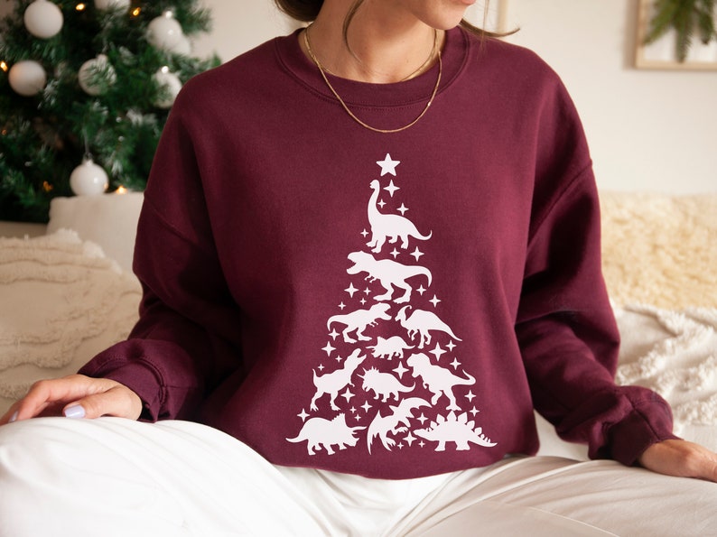 May include: A maroon sweatshirt with a white dinosaur Christmas tree design. The tree is made up of various dinosaurs, including a T-Rex, a stegosaurus, and a brachiosaurus.