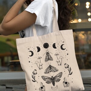 May include: A beige tote bag with a black and white design featuring moons, stars, moths, mushrooms, and crystals.