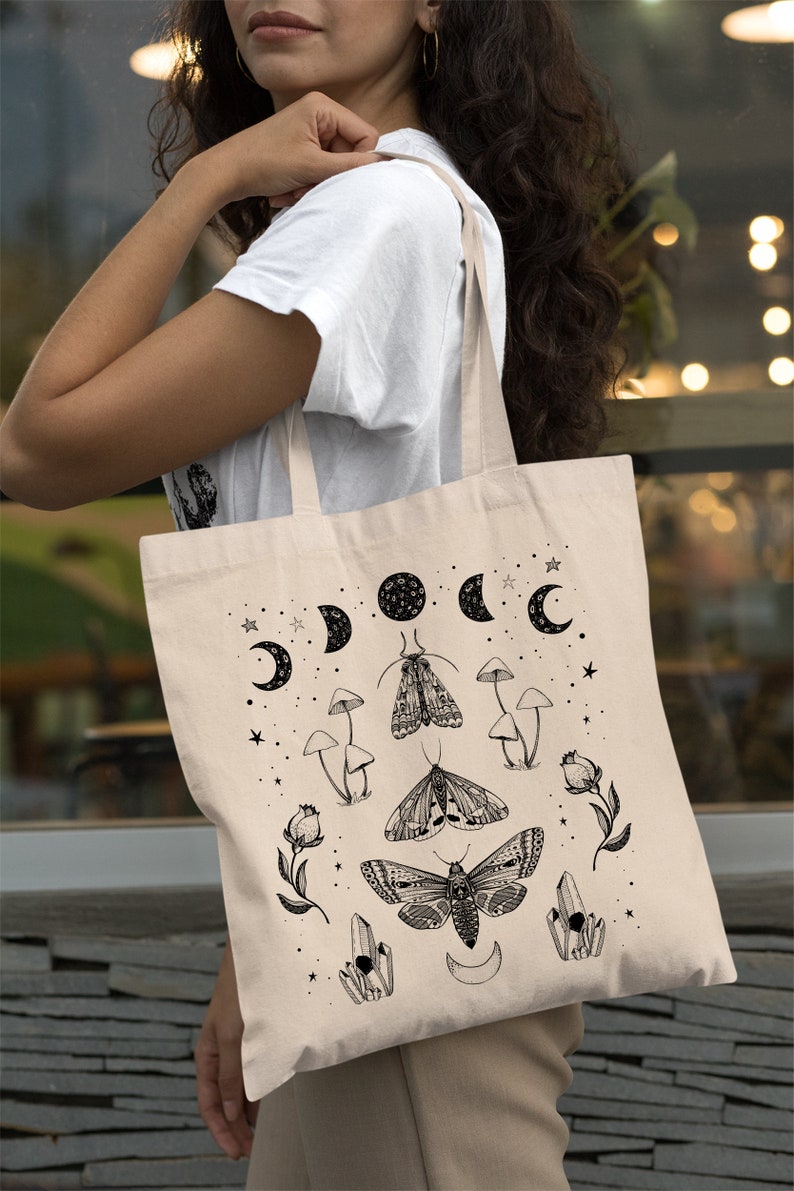 May include: A beige tote bag with a black and white design featuring moons, stars, moths, mushrooms, and crystals.