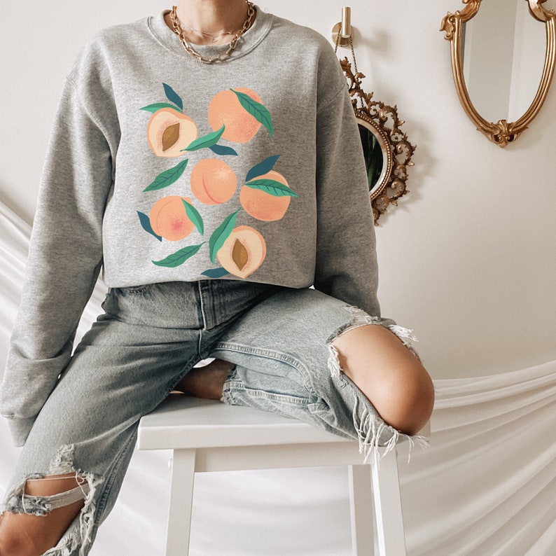 K&ouml;nnte beinhalten: Ein graues Sweatshirt mit einem Pfirsich-Design. Die Pfirsiche sind in verschiedenen Farben illustriert, darunter Rosa, Orange und Gelb. Das Sweatshirt wird von einer Person getragen, die auf einem wei&szlig;en Hocker sitzt.