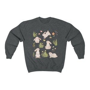 May include: A dark heather grey sweatshirt with a pattern of white bunnies and flowers. The bunnies are in various poses, including sitting, standing, and hopping. The flowers are in shades of pink, yellow, and orange.