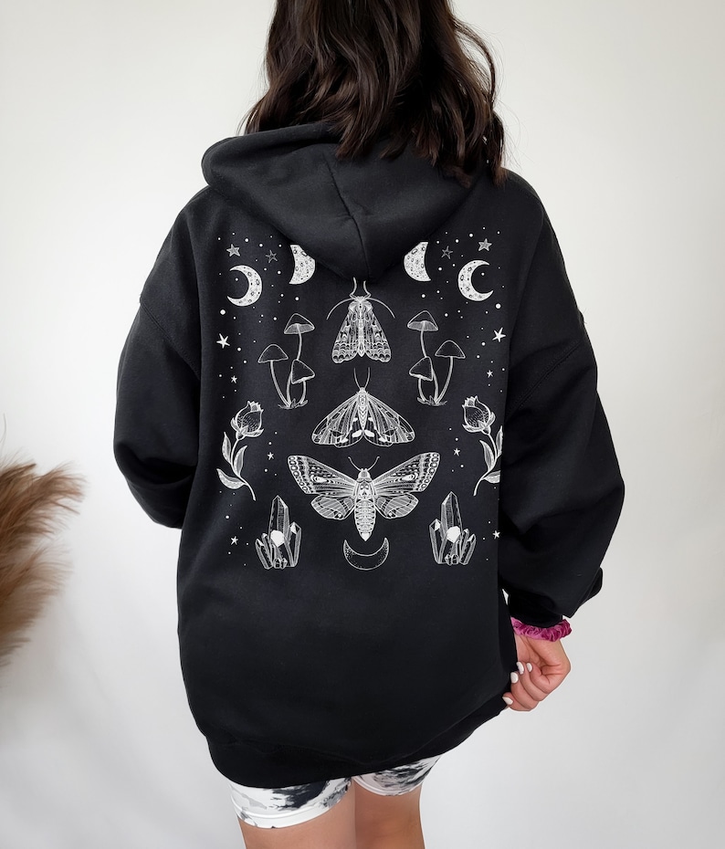 Pu&ograve; includere: Felpa con cappuccio nera con una stampa grafica bianca di falene, cristalli, funghi e lune crescenti.