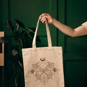 May include: A beige tote bag with a brown butterfly design and geometric patterns. The butterfly has intricate details and is surrounded by crescent moons and other symbols.