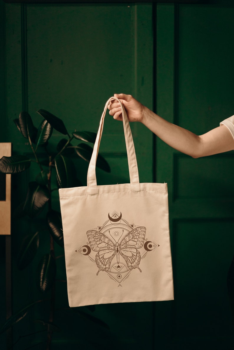 May include: A beige tote bag with a brown butterfly design and geometric patterns. The butterfly has intricate details and is surrounded by crescent moons and other symbols.