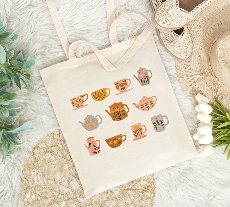 May include: A white canvas tote bag with a floral design featuring teacups and teapots in various colors and patterns. The bag has two long handles.