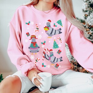 May include: Pink Christmas jumper with a festive design featuring three cartoon pigeons wearing Santa hats and scarves. The jumper is decorated with snowflakes, candy canes, holly, and Christmas trees.