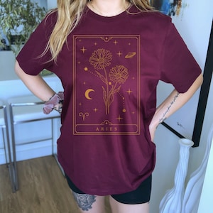 May include: A maroon t-shirt with a gold graphic of a flower with a crescent moon and stars. The graphic is in a rectangular frame with the word "ARIES" at the bottom.