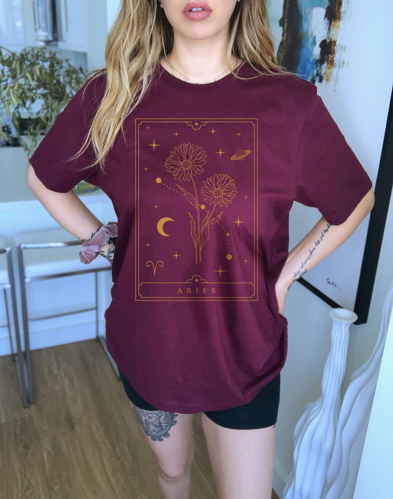 May include: A maroon t-shirt with a gold graphic of a flower with a crescent moon and stars. The graphic is in a rectangular frame with the word "ARIES" at the bottom.