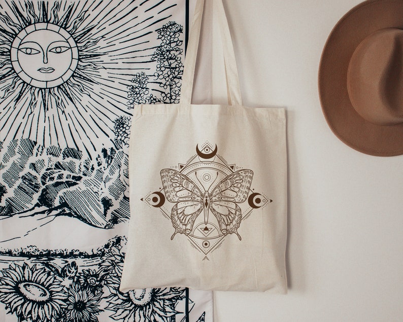 May include: A beige tote bag with a brown butterfly design and geometric patterns. The butterfly is centered on the bag and has a crescent moon on each side.