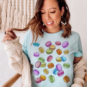 May include: A light blue t-shirt with a colorful macaron pattern. The macarons are scattered across the shirt in a random pattern.