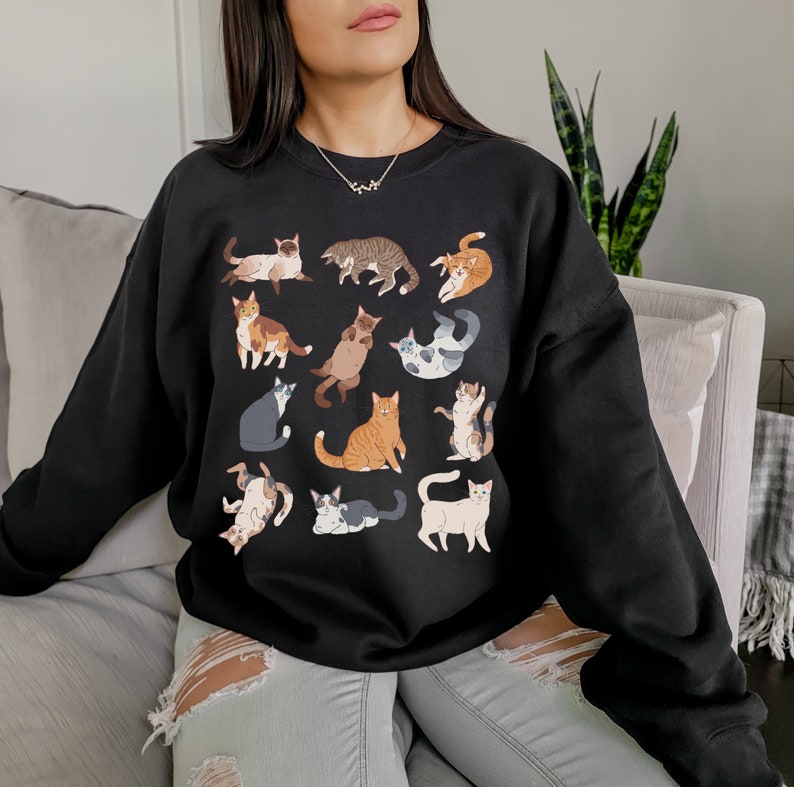 May include: A black sweatshirt with a graphic print of various cats in different poses. The cats are in shades of brown, white, gray, and orange.