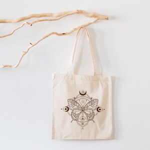 May include: A white canvas tote bag with a brown butterfly design. The butterfly is surrounded by a geometric pattern and crescent moons.