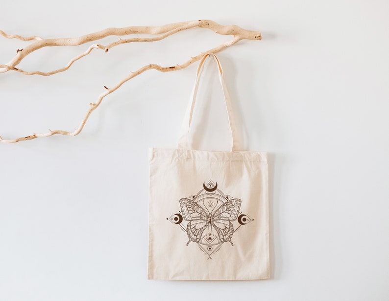 May include: A white canvas tote bag with a brown butterfly design. The butterfly is surrounded by a geometric pattern and crescent moons.