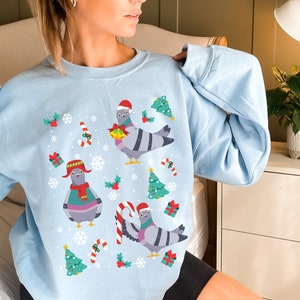 May include: A light blue sweatshirt with a Christmas-themed design featuring cartoon pigeons wearing Santa hats and holding gifts. The design also includes candy canes, snowflakes, and Christmas trees.