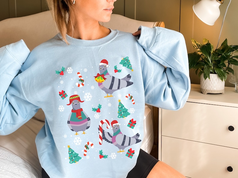 May include: A light blue sweatshirt with a Christmas-themed design featuring cartoon pigeons wearing Santa hats and holding gifts. The design also includes candy canes, snowflakes, and Christmas trees.