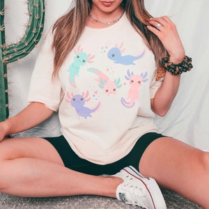 May include: A white t-shirt featuring a graphic of five pink, blue, and purple axolotls with black eyes and smiling faces. The axolotls are surrounded by pink hearts.