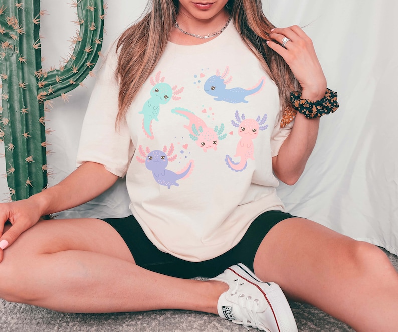 May include: A white t-shirt featuring a graphic of five pink, blue, and purple axolotls with black eyes and smiling faces. The axolotls are surrounded by pink hearts.