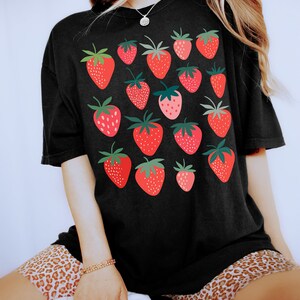Puede incluir: Una camiseta negra de gran tama&ntilde;o con un estampado gr&aacute;fico de fresas rojas. La modelo lleva la camiseta con pantalones cortos de estampado de leopardo.