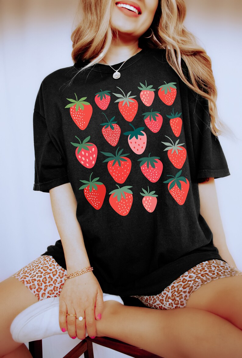 Puede incluir: Una camiseta negra de gran tama&ntilde;o con un estampado gr&aacute;fico de fresas rojas. La modelo lleva la camiseta con pantalones cortos de estampado de leopardo.