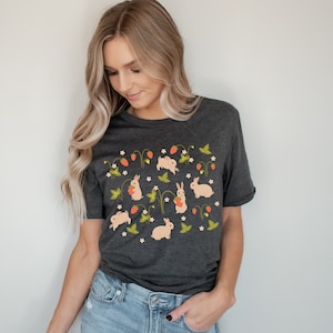 Op de afbeelding: Een donker grijs t-shirt met een patroon van witte konijnen die aardbeien en groene bladeren vasthouden op een witte achtergrond.