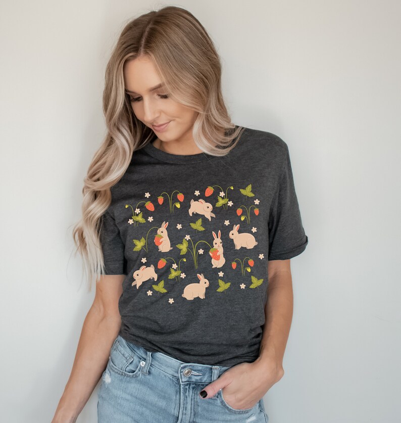 Op de afbeelding: Een donker grijs t-shirt met een patroon van witte konijnen die aardbeien en groene bladeren vasthouden op een witte achtergrond.