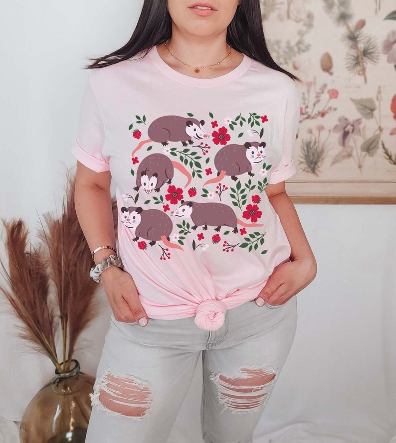 May include: A pink t-shirt with a graphic print of five opossums surrounded by red flowers and green leaves.