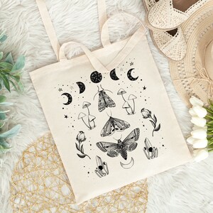 May include: A white canvas tote bag with a black and white design featuring moths, mushrooms, crystals, and crescent moons. The design is reminiscent of a celestial or witchy aesthetic.