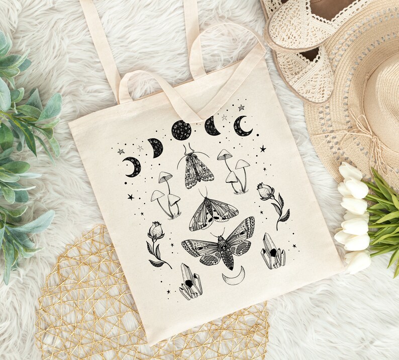 May include: A white canvas tote bag with a black and white design featuring moths, mushrooms, crystals, and crescent moons. The design is reminiscent of a celestial or witchy aesthetic.