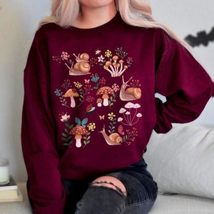 Puede incluir: Una sudadera color burdeos con un estampado gr&aacute;fico colorido de caracoles, setas y flores. El dise&ntilde;o es caprichoso y presenta una variedad de colores, incluyendo rojo, verde, amarillo y azul.