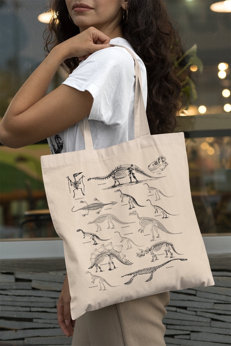 May include: A beige canvas tote bag with black dinosaur skeletons printed on it. The skeletons are arranged in rows and include a variety of dinosaurs, such as a Tyrannosaurus Rex, a Stegosaurus, and a Triceratops.