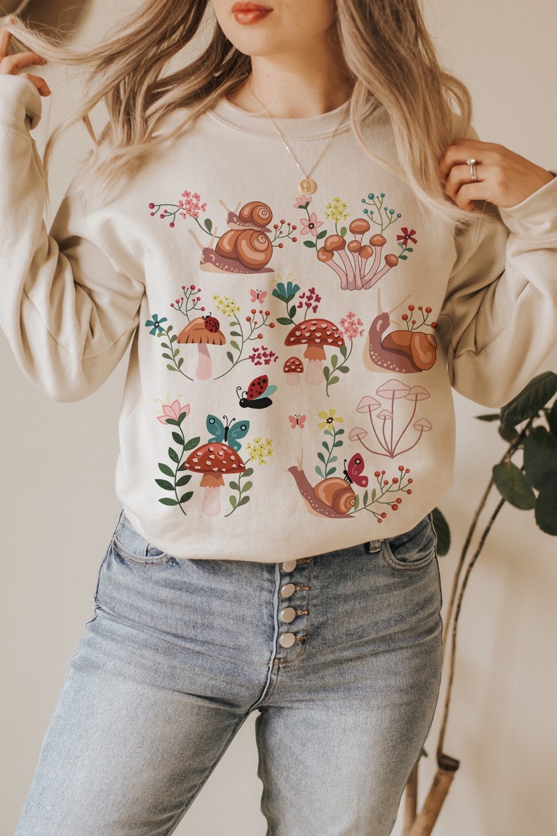 Puede incluir: Una sudadera beige con un dise&ntilde;o colorido de setas y caracoles. El dise&ntilde;o incluye setas rojas y blancas, caracoles marrones, hojas verdes y mariposas de colores.