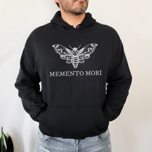K&ouml;nnte beinhalten: Schwarzer Kapuzenpullover mit einem wei&szlig;en Totenkopf-Motten-Motiv und dem Text "MEMENTO MORI" auf der Vorderseite.